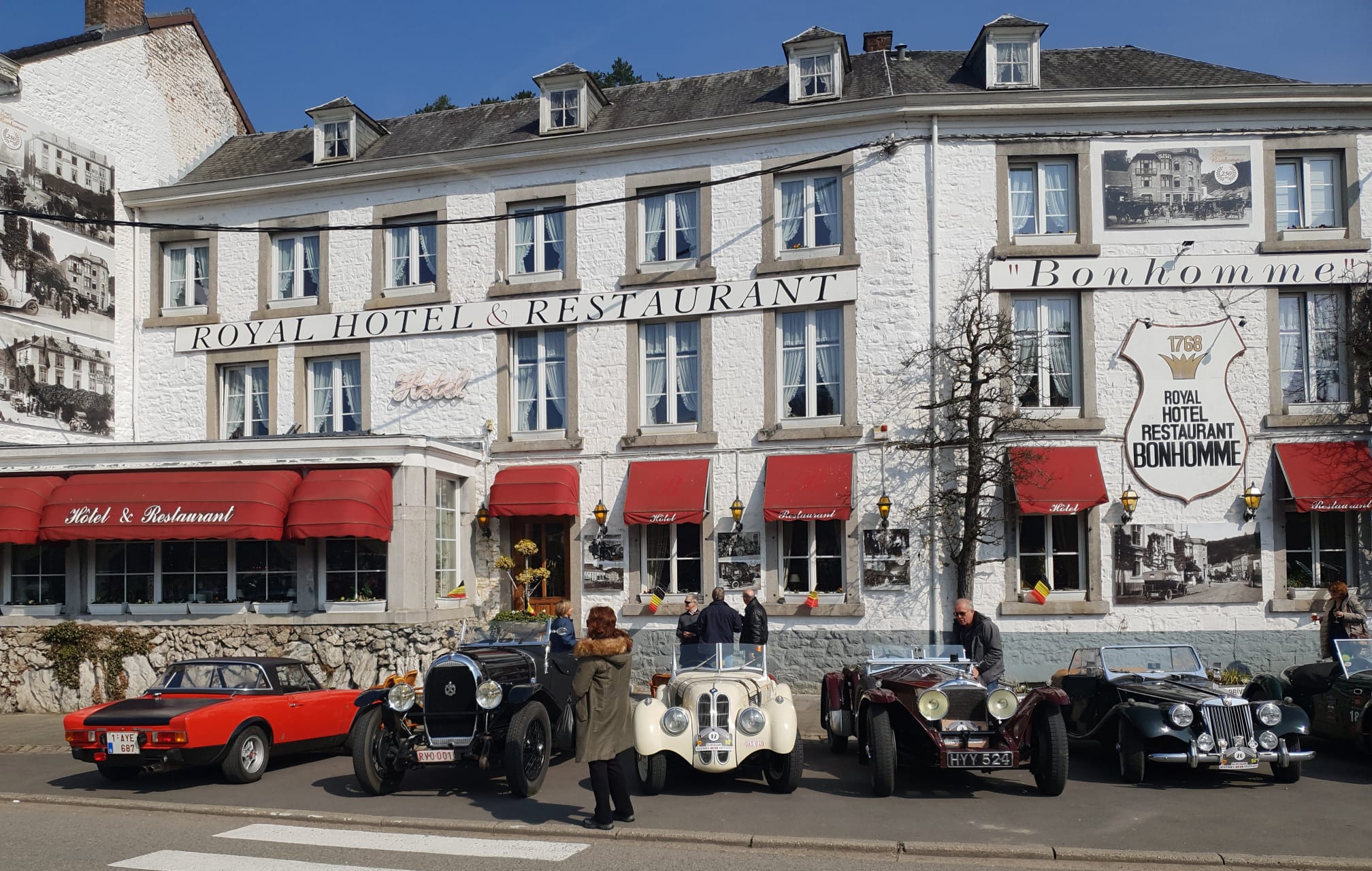 Façade du Royal Hôtel Restaurant Bonhomme avec voitures anciennes, auvents rouges, ciel bleu